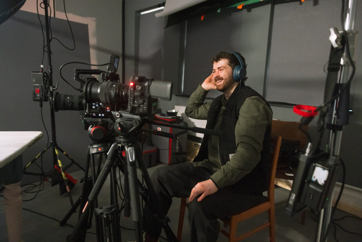 In a video production studio and man wearing headphones and sitting behind a studio video camera is smiling and has one hand on his headphones